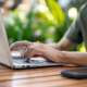 Close up of a man typing on laptop with both hands while sitting at wooden table with phone aside