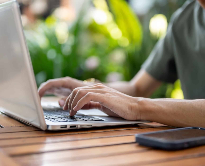 Close Up Of A Man Typing On Laptop With Both Hands While Sitting At Wooden Table With Phone Aside
